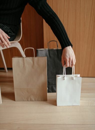 From above of crop faceless male putting various paper bags on wooden floor while sitting on chair at home after shopping